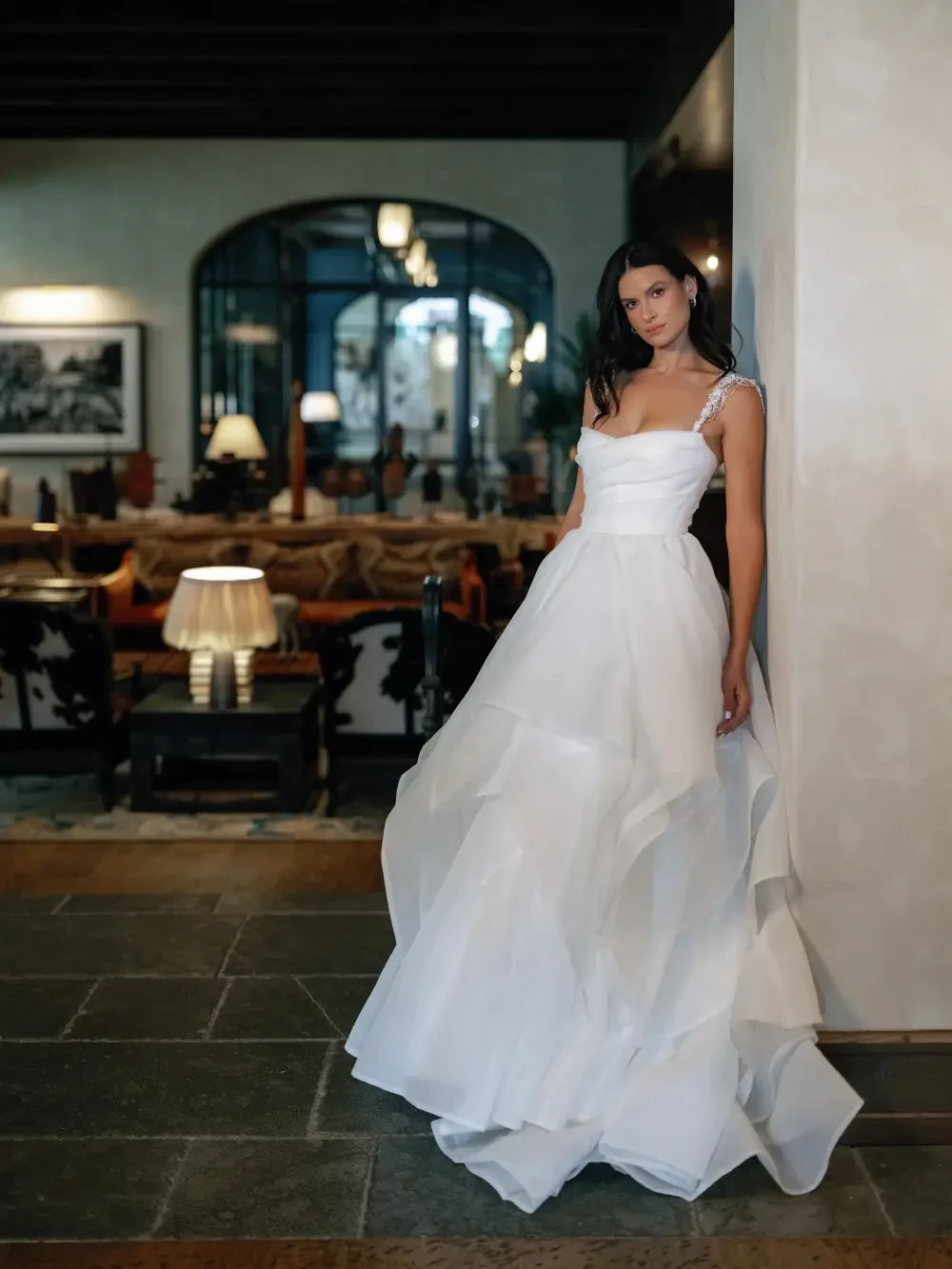 A woman in a flowing white dress stands near a wall in a stylishly decorated interior space.