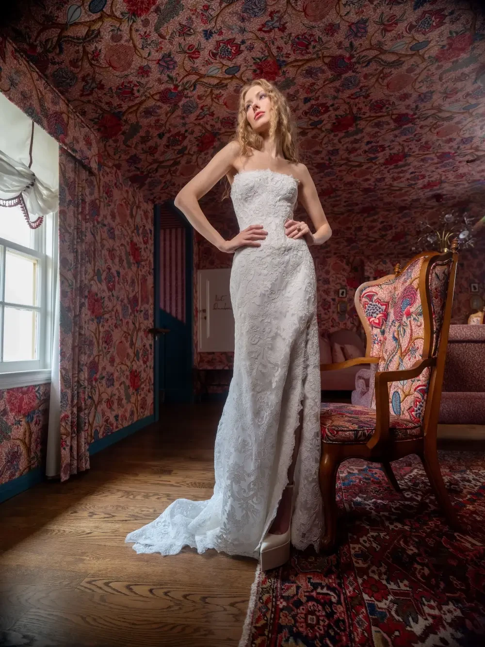 A model wearing a white lace wedding dress stands in a room with floral wallpaper and Victorian-style furniture.