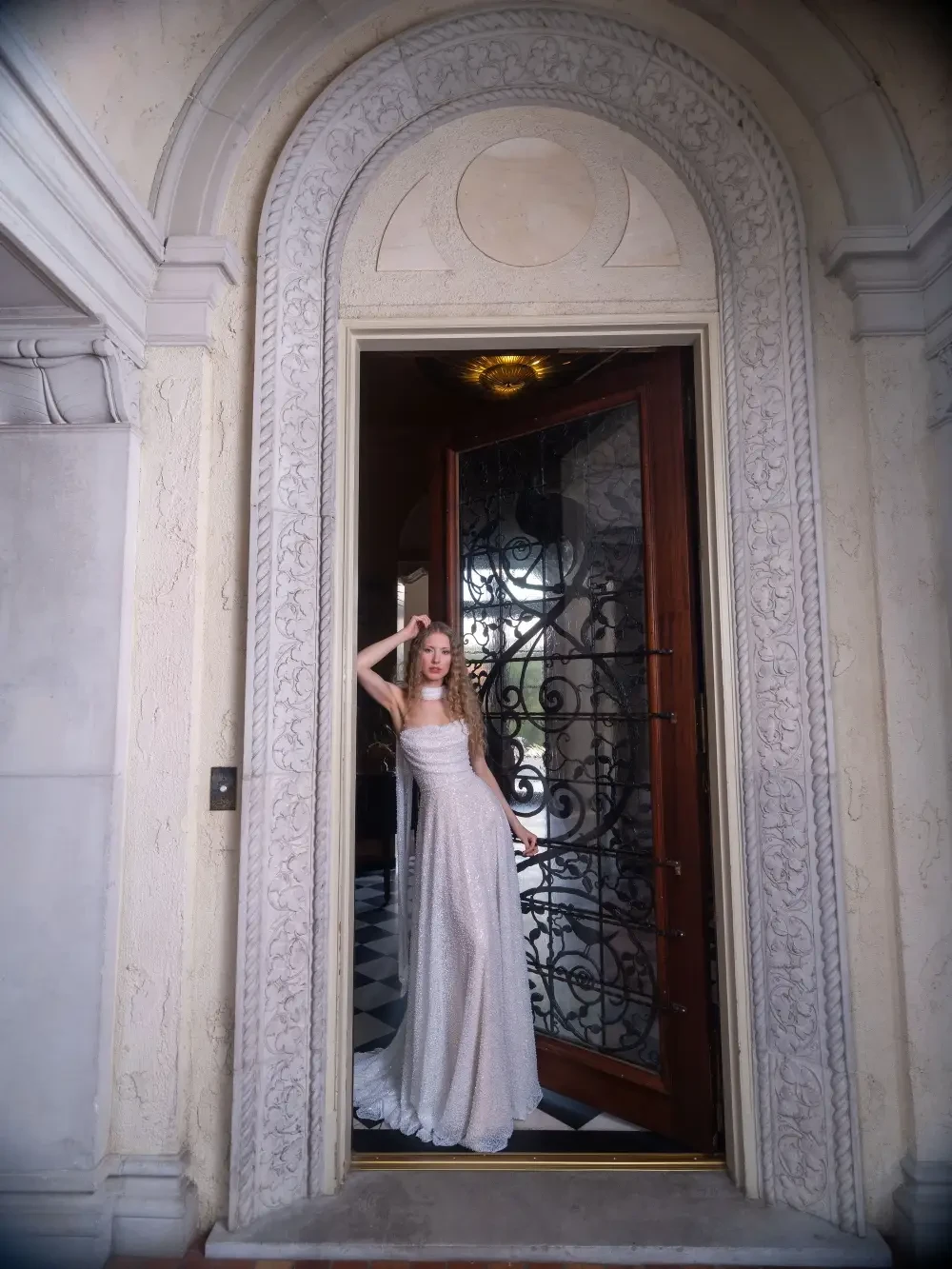 A woman in a flowing dress stands in an ornate doorway, framed by intricate architectural details.