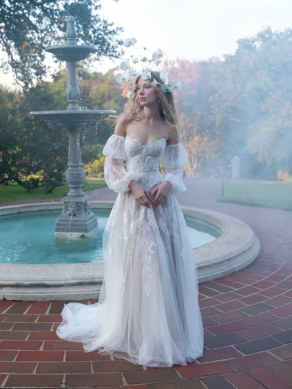 A woman in a floral crown stands by a fountain, wearing an elegant, flowing white gown in a garden setting with soft mist.