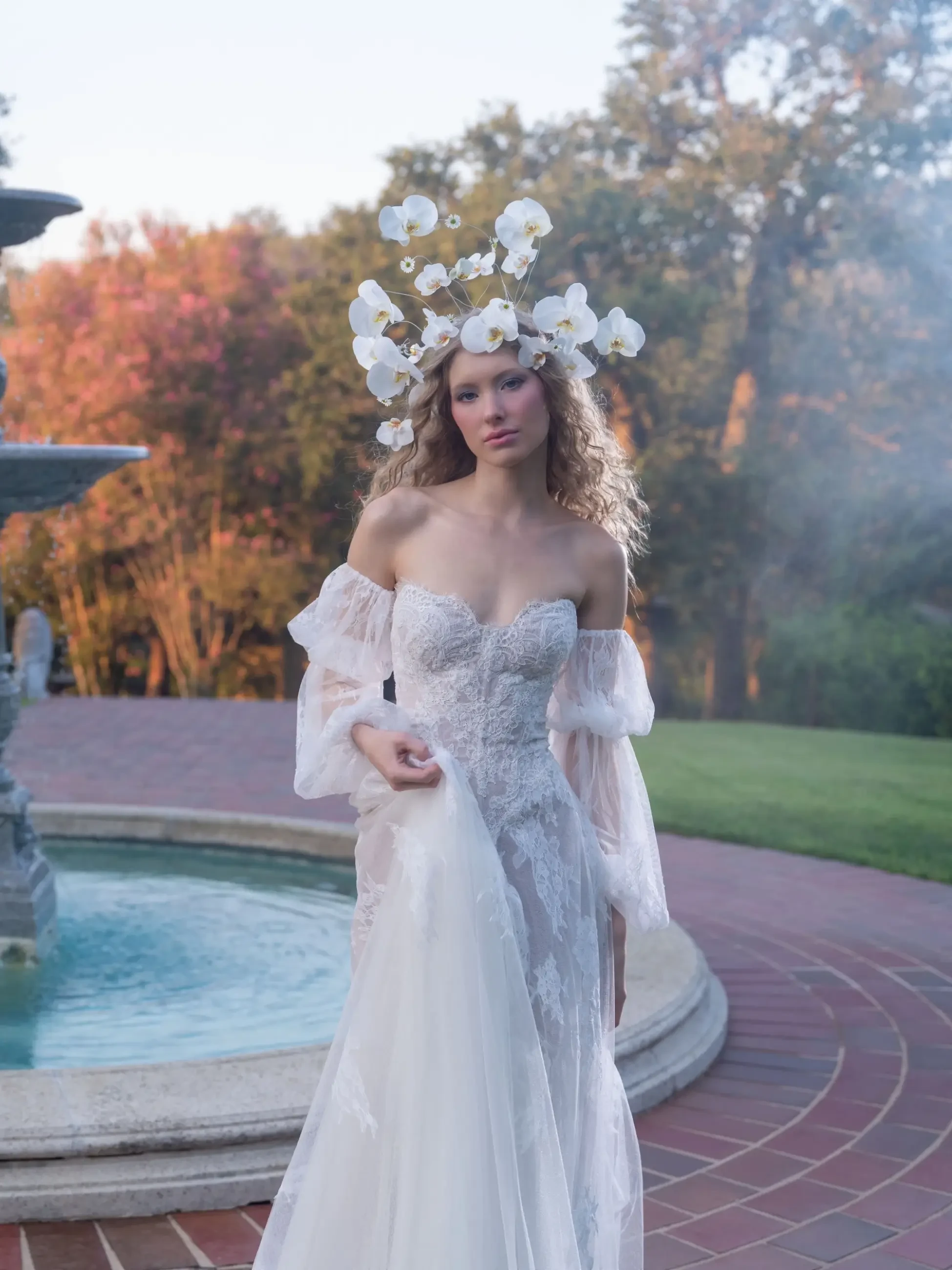 A model wearing a white wedding dress stands near a fountain, adorned with a floral crown made of orchids. Soft lighting and natural scenery create an ethereal atmosphere.