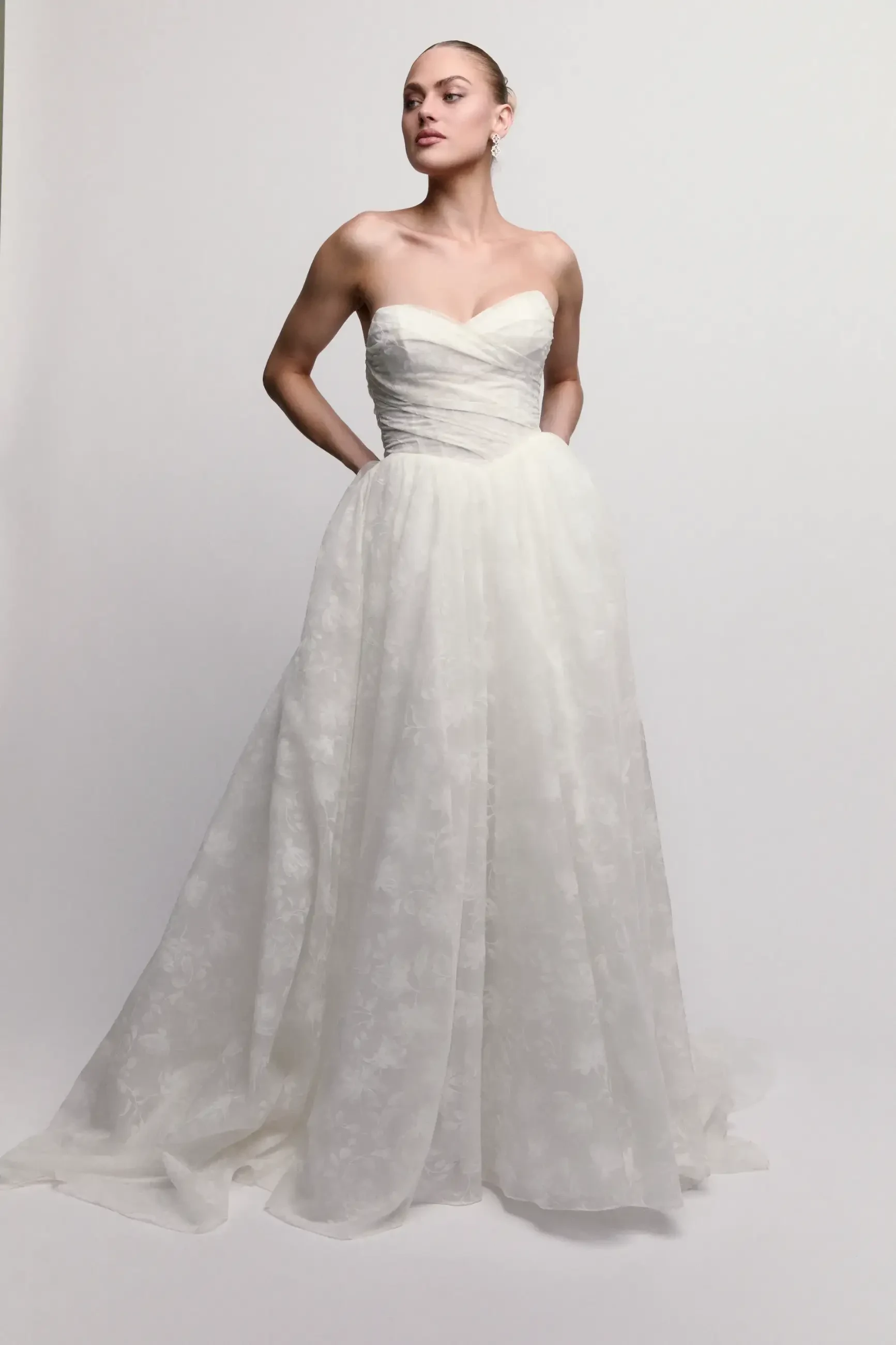 A woman poses gracefully in a strapless white wedding gown with a fitted bodice and flowing skirt, exuding elegance and poise against a plain backdrop.