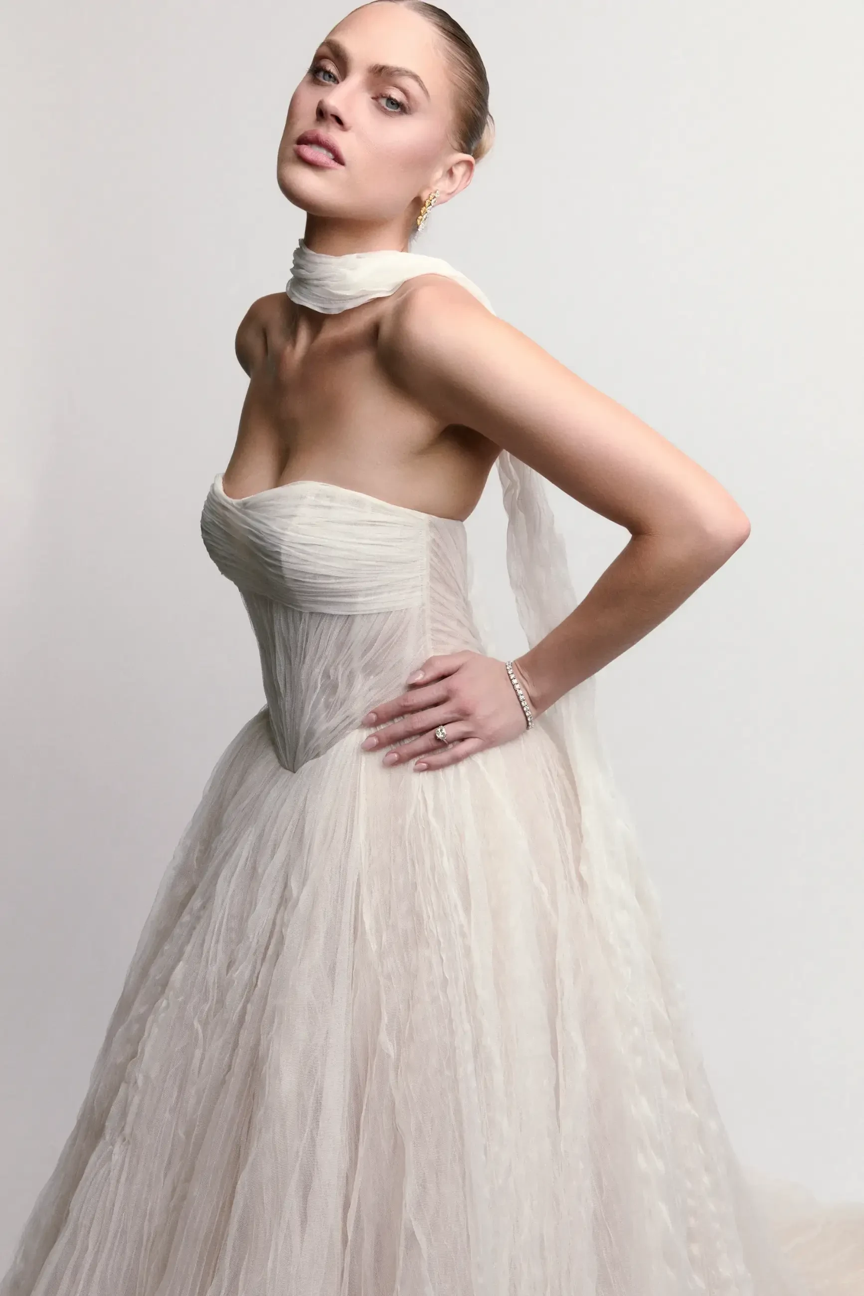 A model in a strapless, light-colored gown with a fitted bodice and flowing skirt, posing elegantly with one hand on her hip.