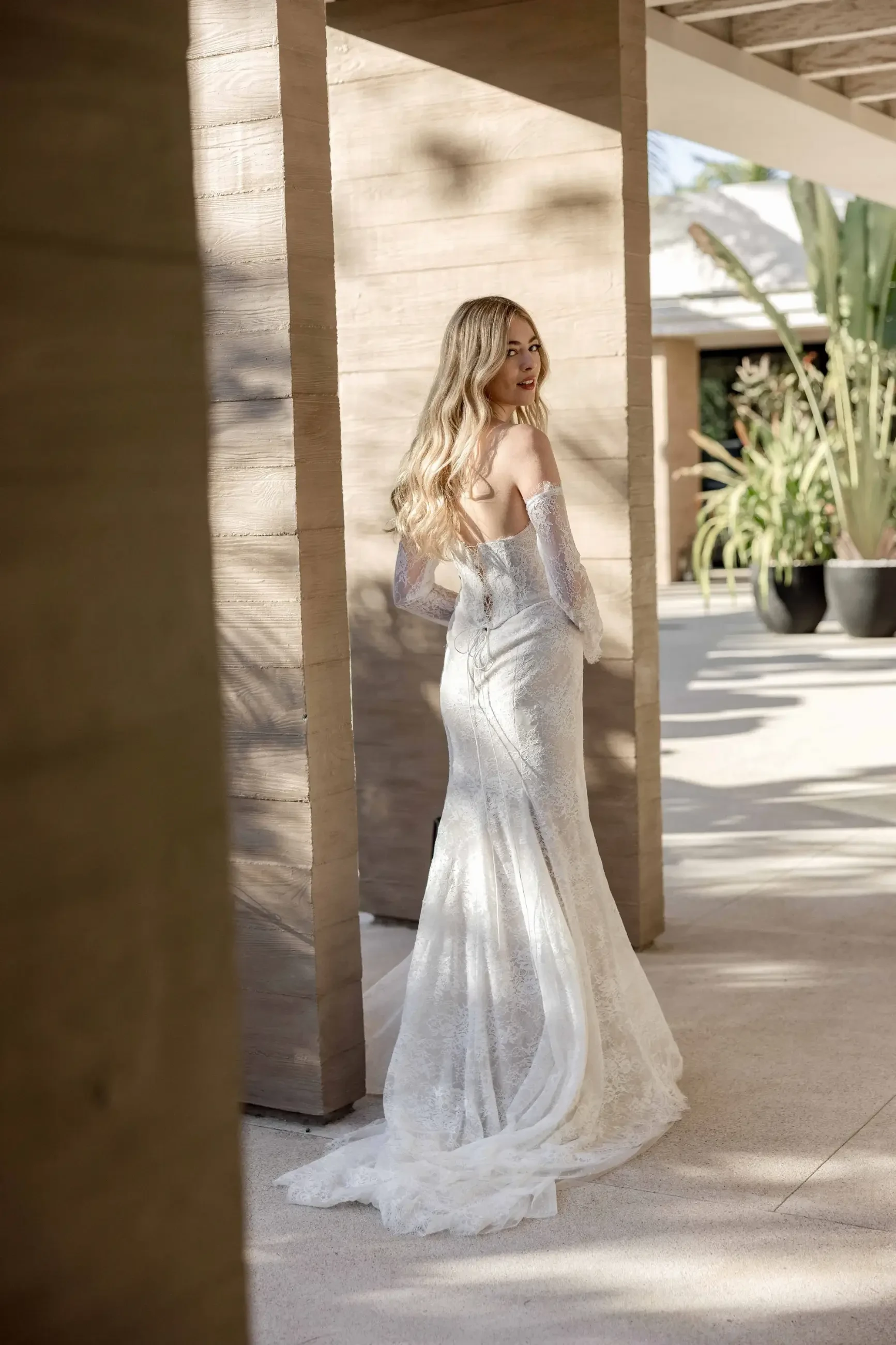 A woman in a white wedding dress poses in an outdoor setting, standing with her back to the camera and looking over her shoulder. The surroundings include architectural elements and lush greenery.