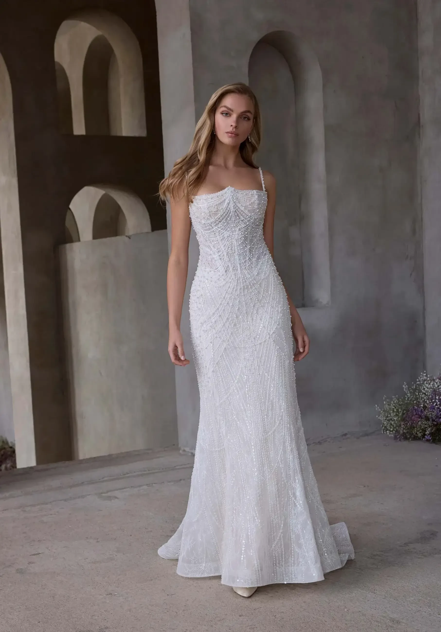 Model is wearing a fit-and-flare wedding dress with an angel wing neckline, thin straps, basque waist, and pearl-embroidered tulle.