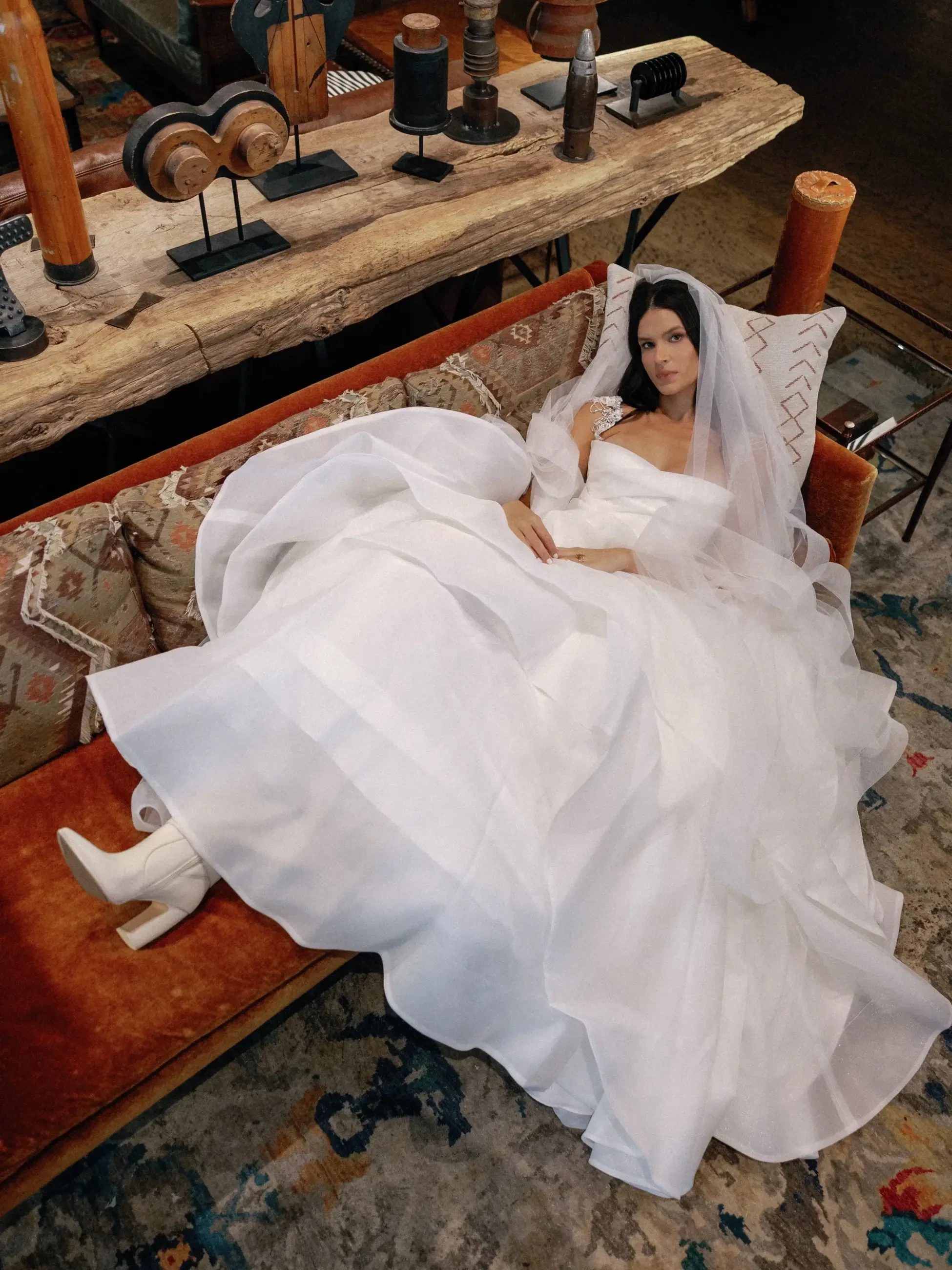 A woman in a flowing white wedding dress and veil reclines on a vintage sofa. Surrounding her are rustic wooden decor and an ornate patterned rug.