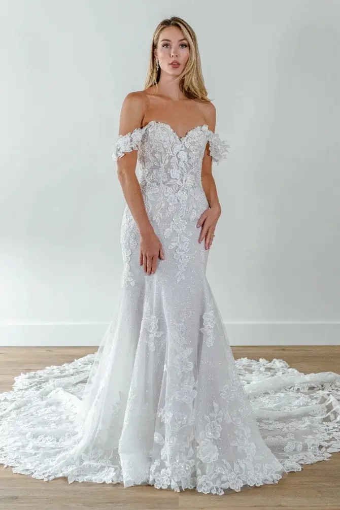 Model is wearing an a-line bridal gown with glitter tulle and off the shoulder straps