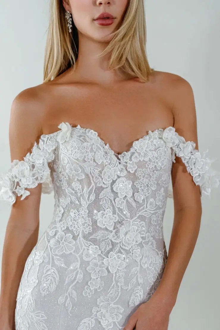 Model is wearing an a-line bridal gown with glitter tulle and off the shoulder straps