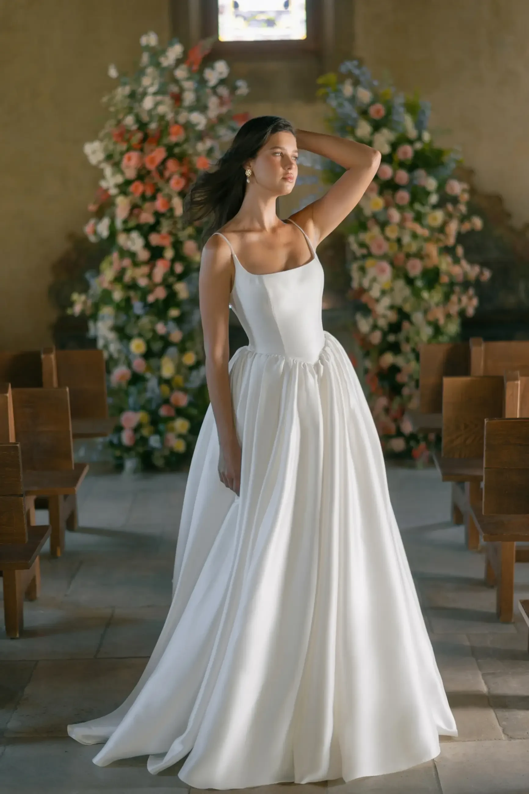 A woman in a flowing white wedding gown stands in a church, surrounded by colorful floral arrangements. She exudes elegance and serenity.