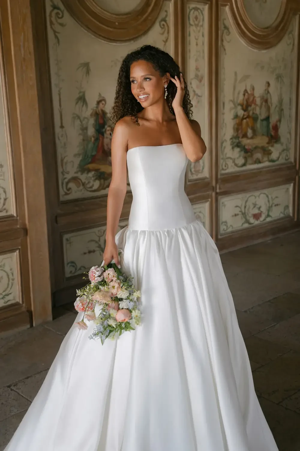 A woman in a strapless white wedding dress holding a bouquet of flowers, standing in an ornate room.