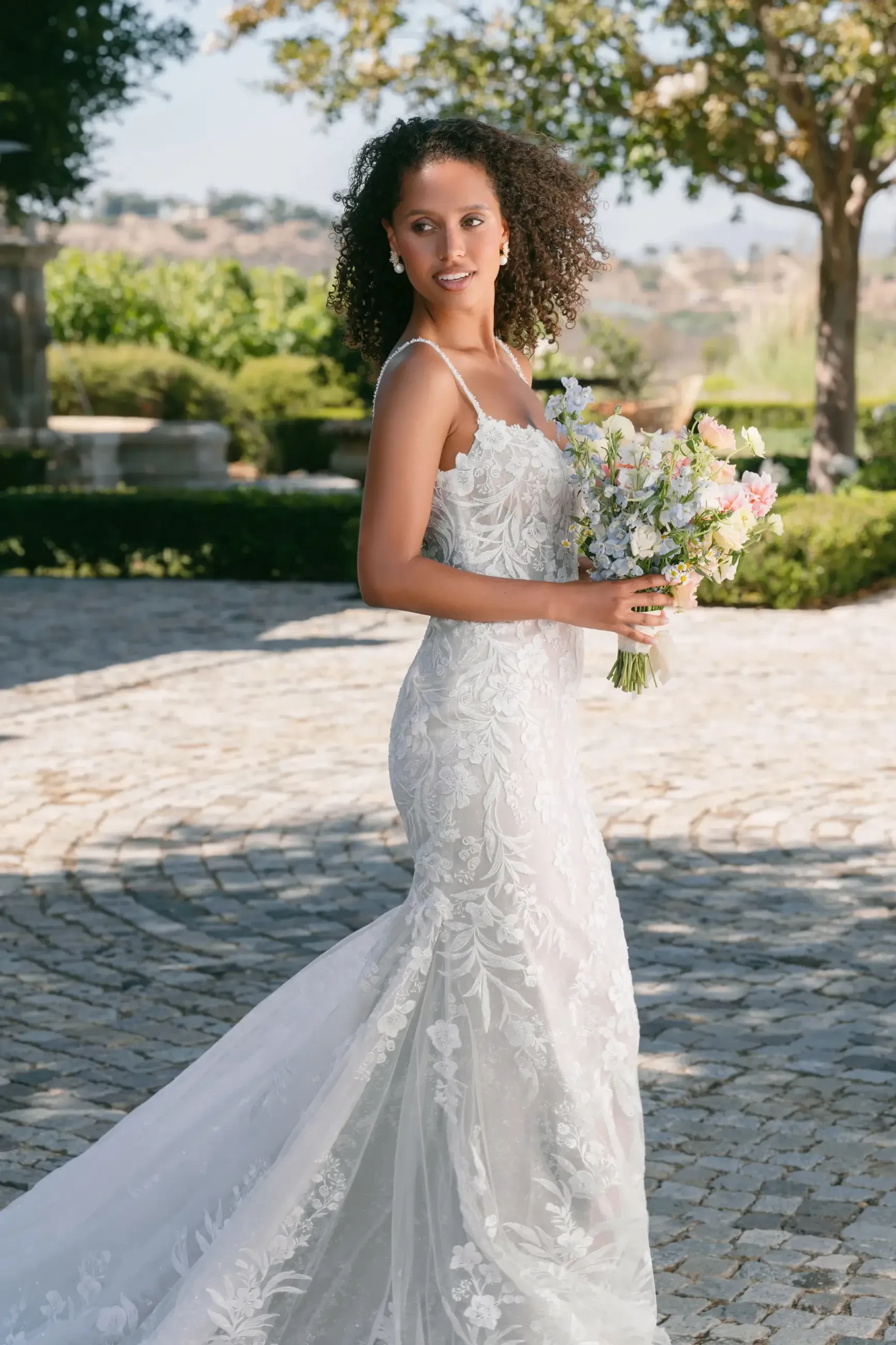 A woman wearing a wedding dress stands outdoors, holding a bouquet of flowers, with greenery and stone pathways in the background.