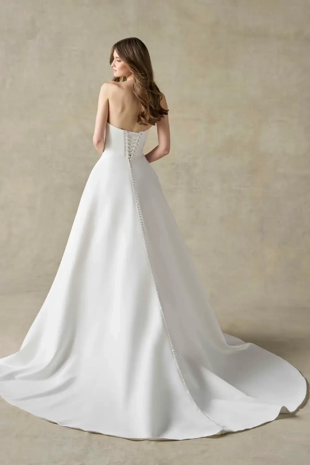 A woman wearing a strapless white wedding gown, viewed from the back, with a long train and buttons along the seam.
