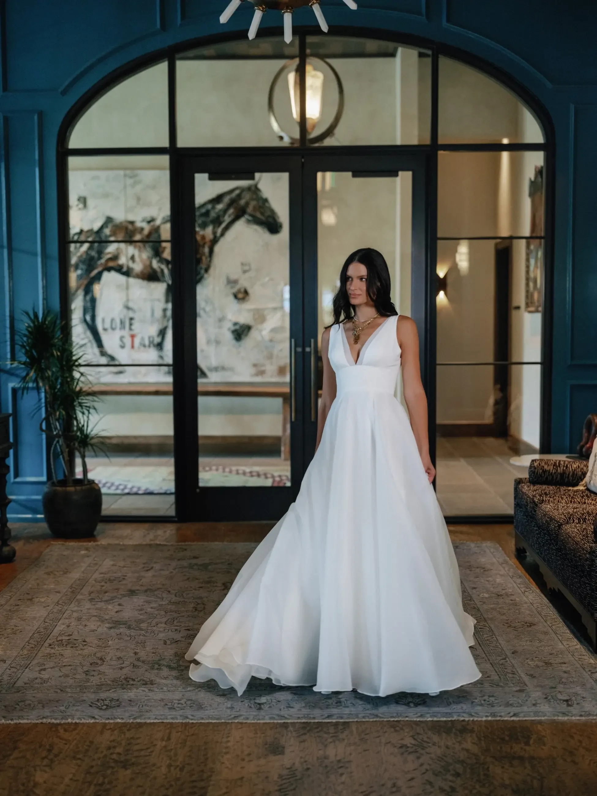 A woman in a white wedding dress stands in a beautifully decorated room with large windows and artwork.