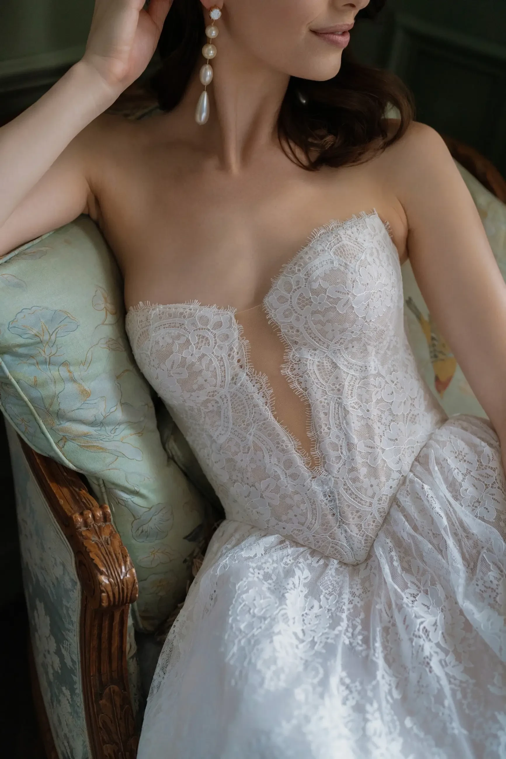 A close-up of a woman wearing a strapless wedding dress with intricate lace detailing, seated on an ornate chair.