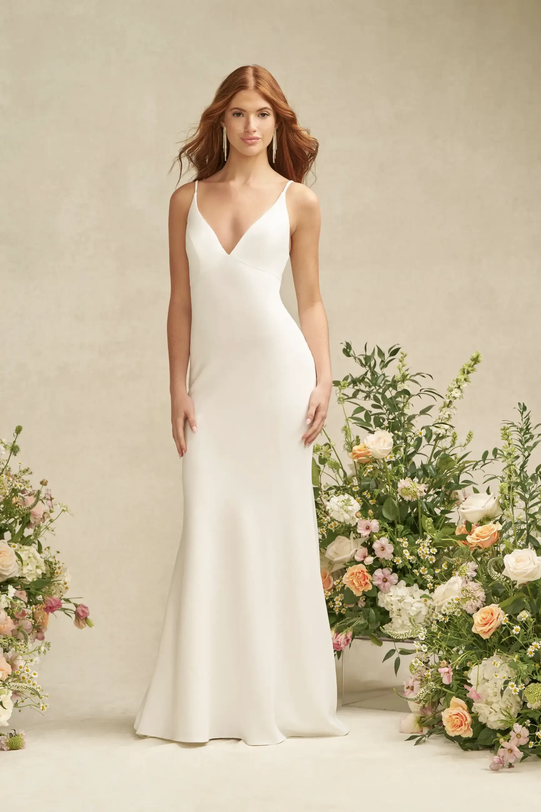 A model wearing a sleek white dress stands in front of floral arrangements in soft pastel colors.