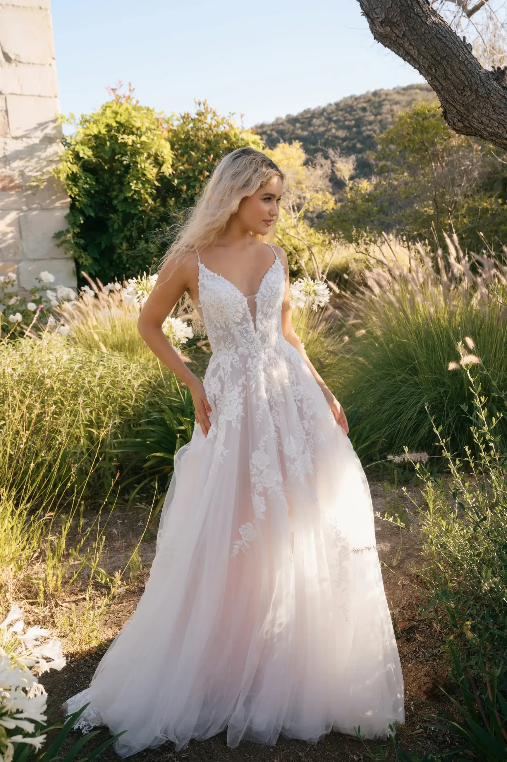 A woman in a flowing wedding gown stands in an outdoor setting with greenery and sunlight.