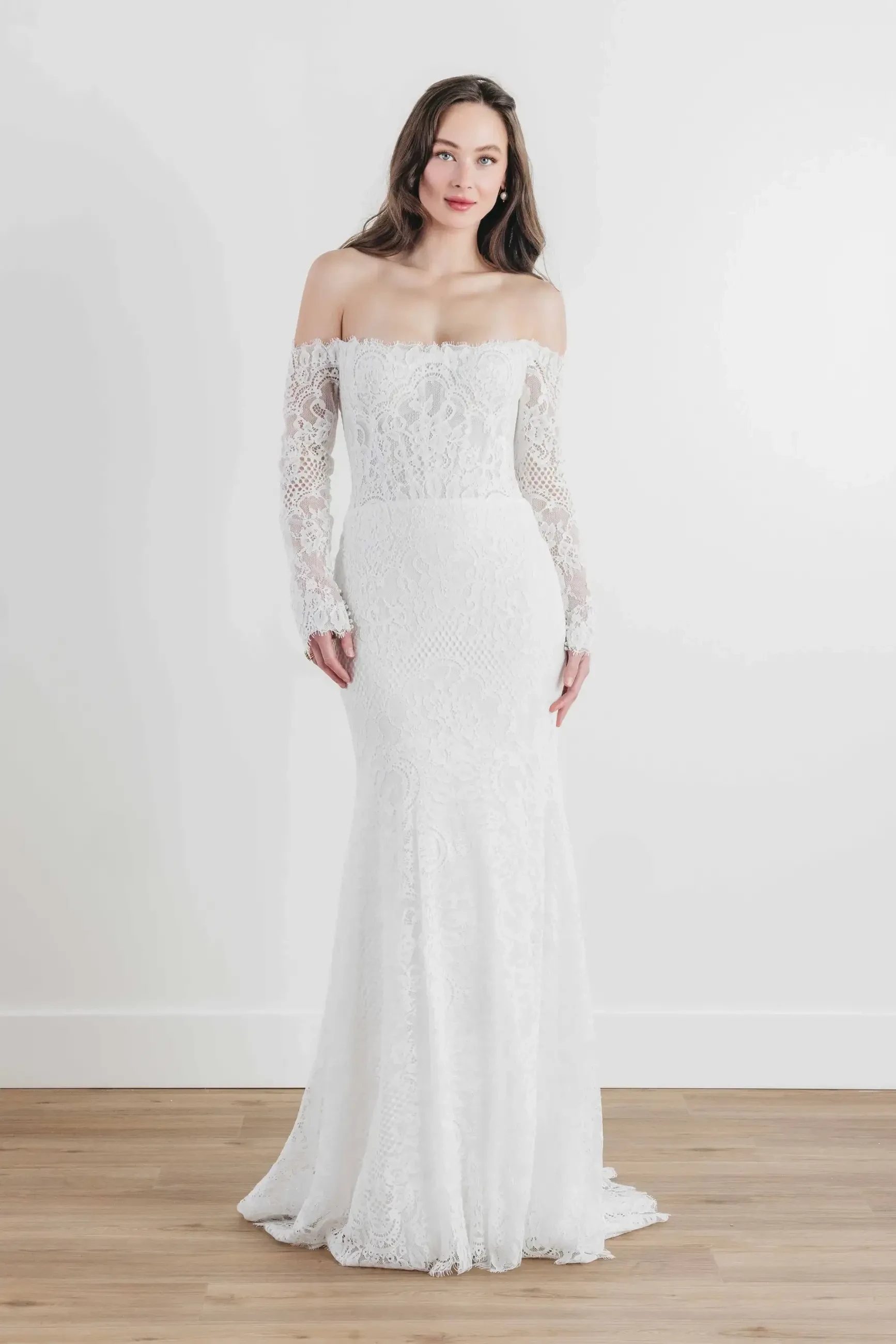 Model is wearing a fitted lace gown with long sleeves, an off-the-shoulder neckline, and an illusion low back.