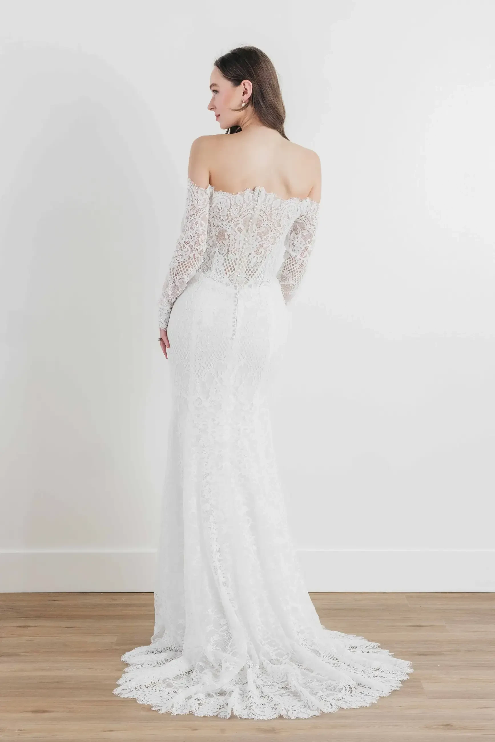 Model is wearing a fitted lace gown with long sleeves, an off-the-shoulder neckline, and an illusion low back.