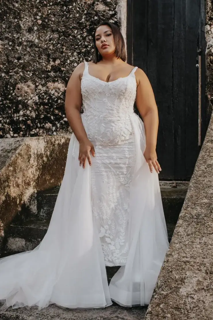 A woman in a wedding dress stands confidently on a staircase, with a textured stone wall and dark door behind her. The dress features intricate lace details and a flowing train.