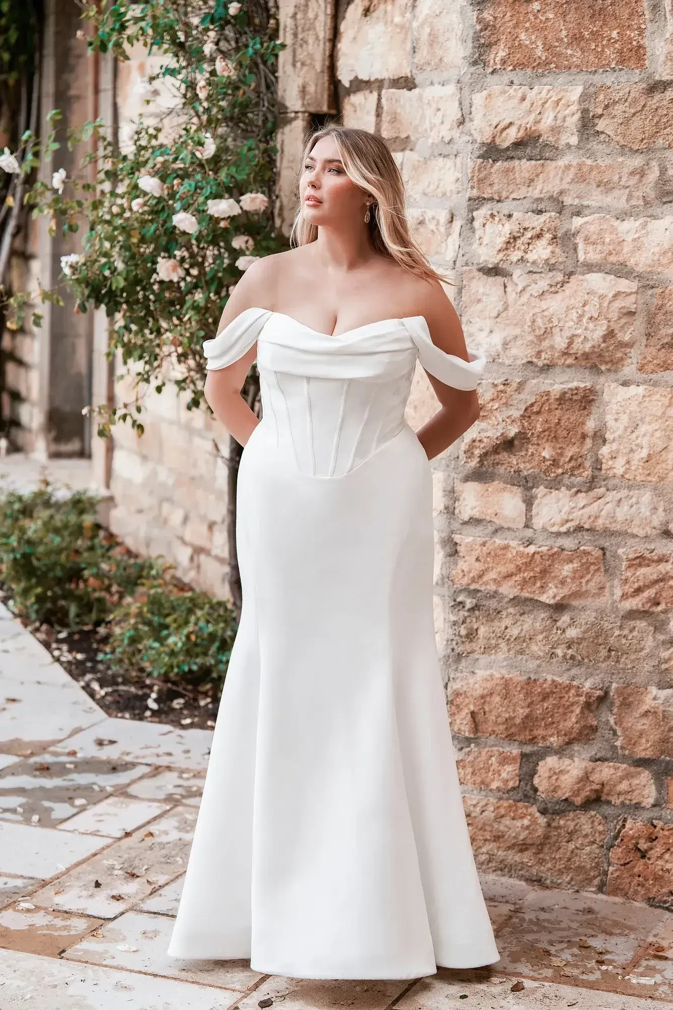 A woman wearing a white off-the-shoulder gown stands outdoors near a stone wall adorned with greenery and flowers.