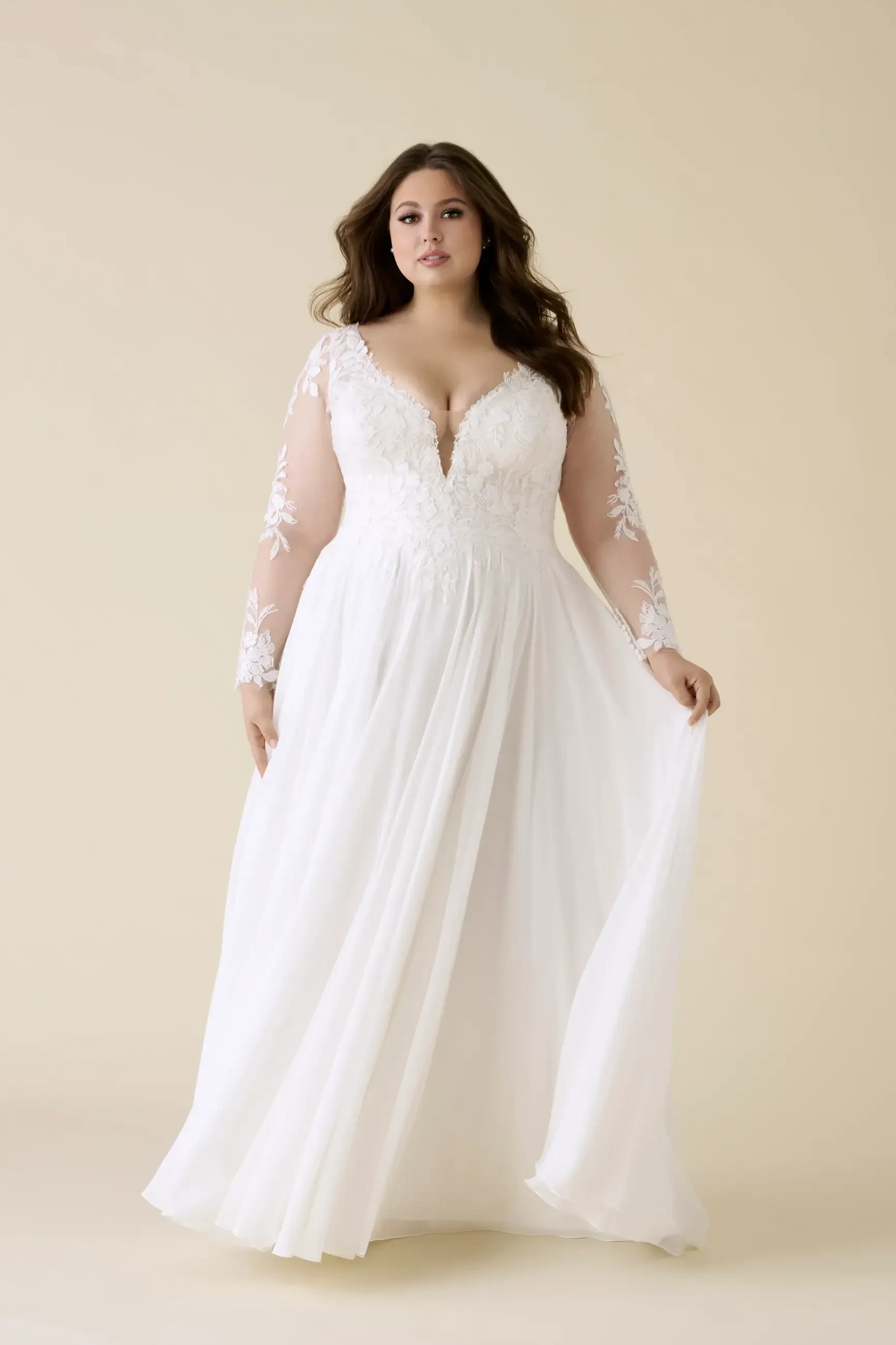 A plus-size woman wearing a white wedding dress with long sleeves and a deep neckline, standing against a light beige background.